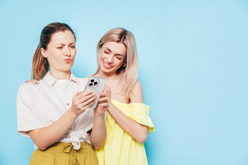 Two young beautiful smiling brunette hipster female in trendy summer clothes. Carefree women posing near blue wall. Positive models having fun. Cheerful and happy. Holding smartphone. Using apps
