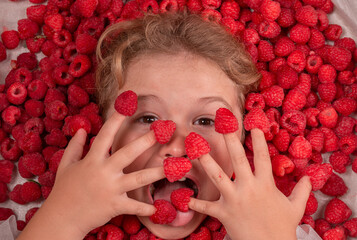 Funny kids face near raspberry background. Cute child eats raspberries from fingers. Fresh raw...