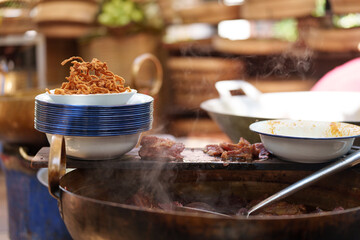 Crispy fried onions are served in a stack of bowls next to a steaming hot pot with sliced meat at an asian street food stall, creating an authentic culinary scene
