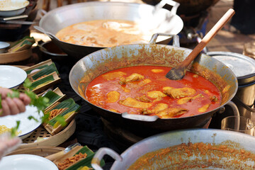Large woks simmering with malaysian chicken rendang, a rich and flavorful coconut milk-based curry, are being prepared at an outdoor market, alongside traditional garnishes and accompaniments