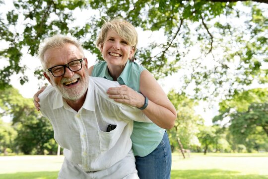 Elderly couple enjoying a sunny day in park. Both are smiling and appear happy. The man wears glasses. They are surrounded by lush green trees. Elderly couple hugging, enjoying the outdoors in park.