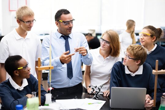 Diverse students and teacher in a science lab. Group of students learning chemistry. Teacher explaining experiment. Students wearing safety goggles. Teacher teaching a diverse high school class. - Powered by Adobe