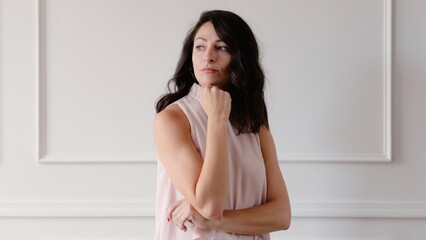 Woman in a sleeveless top poses thoughtfully against a white wall. She has long dark hair and a contemplative expression, exuding elegance and poise. Beautiful mature woman.