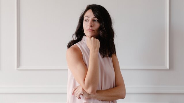 Woman in a sleeveless top poses thoughtfully against a white wall. She has long dark hair and a contemplative expression, exuding elegance and poise. Beautiful mature woman.