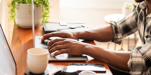 Person typing on a laptop at a wooden desk. Hands on keyboard, coffee mug nearby. Working on a laptop, focused on typing. Cozy workspace with a laptop. African American man working on laptop.