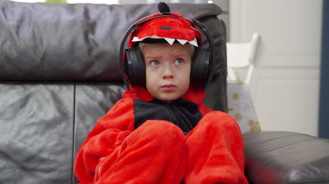 Child wearing red dragon costume in bluetooth wireless large over ear headphones. Little boy in animal kigurumi pajamas playing on video game console at home. Concentrated face of child.