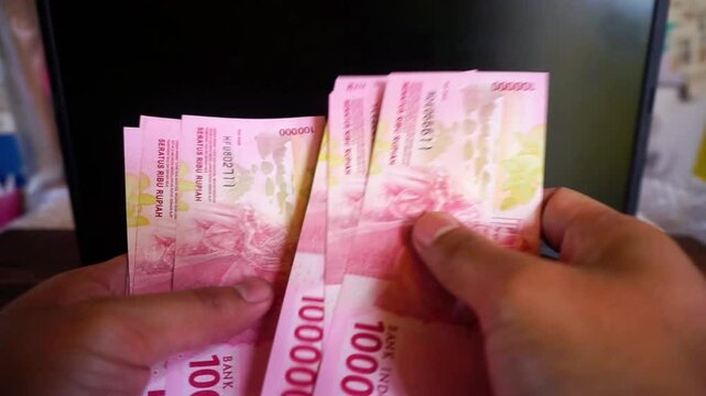 Asian man counting income of hundred thousand rupiah Indonesian currency in front of laptop