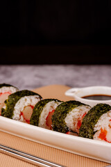 Maki sushi roll with salmon, crab stick, soy sauce, and metallic chopsticks over a brown mat. Black background