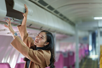 Young woman adjusting an overhead compartment in an airplane cabin. She is smiling and appears to...