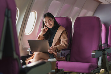 Asian businesswoman smiling and working on her laptop while wearing earbuds during a flight on an...