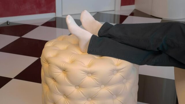 A man takes the black remote control from a white leather round ottoman, sits down and puts his feet on it and turns on electronic equipment (TV, music center, satellite broadcast)