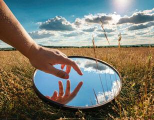 nature concept - hand touching sky reflection in round mirror on summer field 