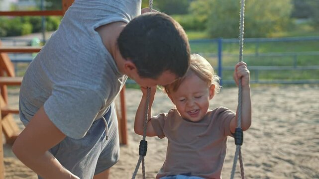 Father play with son at playground. Son enjoys swing ride while father pushes. Happy bonding moment between father and son outdoors. Family play at sunny playground. Smiling son swing with father.