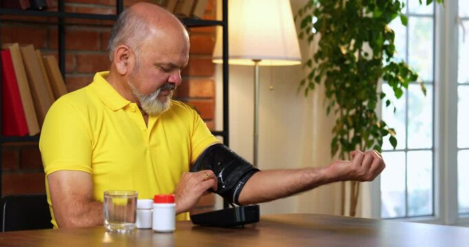 Indian asian senior man checks blood pressure at home, sees high reading, and looks distressed while holding medication bottle