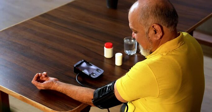 Indian senior man checks blood pressure at home with a digital monitor, sees normal reading, and smiles while holding a pill bottle for routine health care