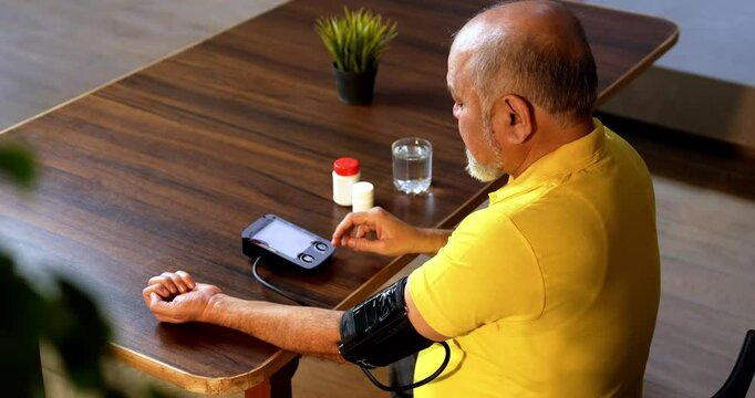 Indian asian senior man checks blood pressure at home, sees high reading, and looks distressed while holding medication bottle
