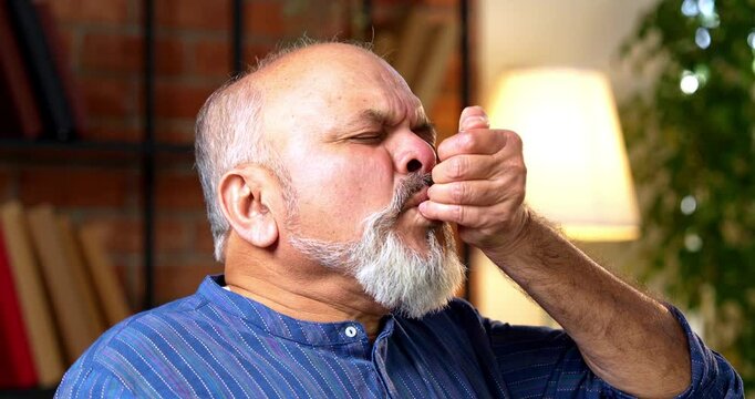 Indian senior retired man feels breathless from an asthma attack, uses inhaler to regain breath while sitting at his home desk