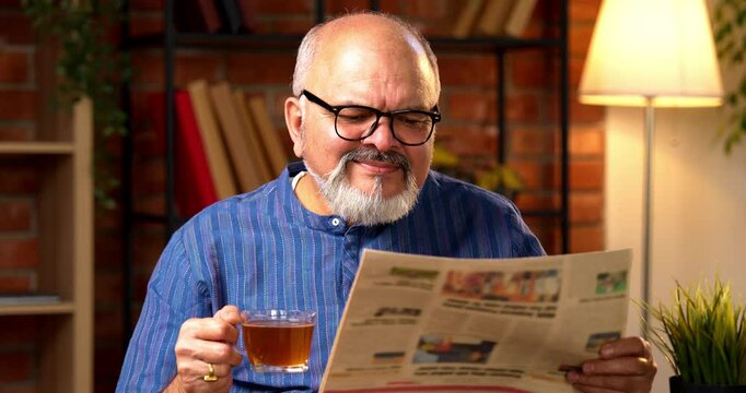 Indian senior retired man wears kurta, reads morning newspaper while sipping black coffee at home desk, staying updated on current business affairs and feeling relaxed