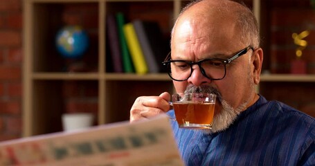 Indian senior retired man wears kurta, reads morning newspaper while sipping black coffee at home desk, staying updated on current business affairs and feeling relaxed
