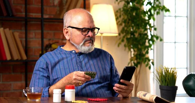 Indian senior retired man in kurta checks medicine prescription on smartphone with pill bottle and medicine strips, ensuring correct dosage