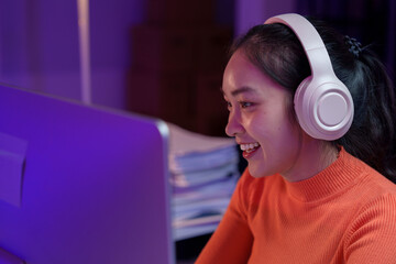 Focused young woman wearing headphones, working on a computer in a dimly lit room. Stacks of papers...