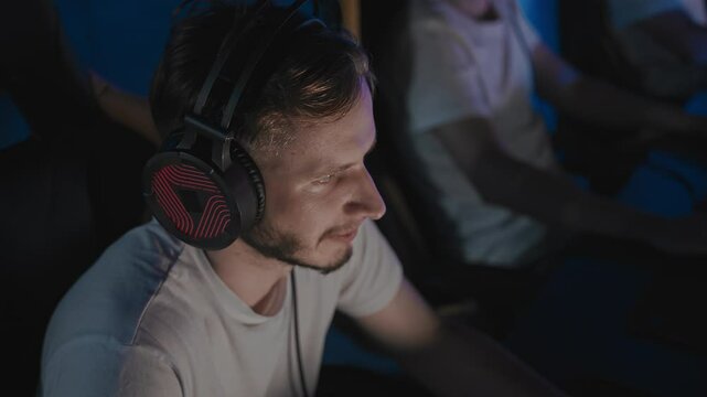 Adult bristly guy chews gum and plays real-time strategy game on PC at gaming cafe during Esports event. Caucasian gamer turns his head to stretch neck. Man playing video game on desktop computer