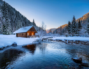 Small wooden cabin glowing on a river in the winter mountains 