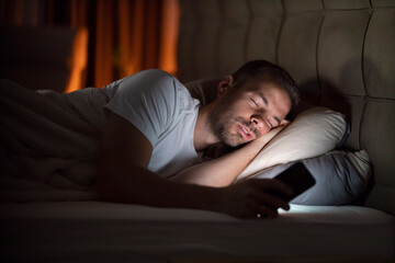 A man is sleeping in bed in dark room with cellphone in hands illuminated by phone light.