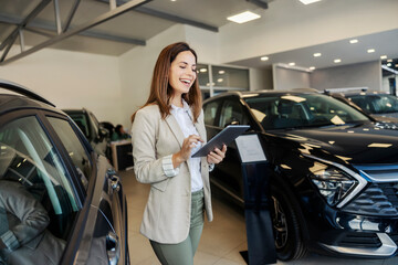 Happy female car seller scrolling on tablet at classy car showroom.