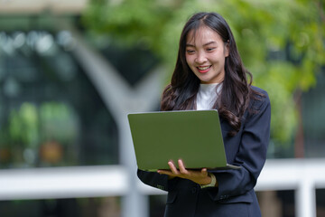 Fototapeta premium Young asian businesswoman is smiling while using a laptop outdoors, enjoying the fresh air and greenery of an urban park, suggesting a blend of work and leisure