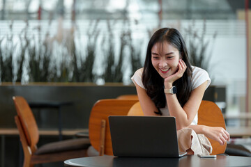 Naklejka premium Smiling young woman using a laptop at a cafe, showcasing remote work or online learning. Modern setting with natural light and comfortable seating, highlighting digital connectivity