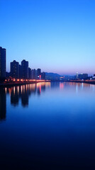 Obraz premium Serene twilight cityscape reflected in calm waters; modern architecture, city lights illuminate the tranquil river at dusk.