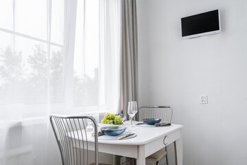 simple kitchen in white tones, plates and glasses on kitchen table, platter of fruits. table near window. TV on wall.