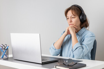 Serious freelance professional woman wearing big headphones listening and watching video, online conference, video meeting, webinar or studying at the web course on laptop