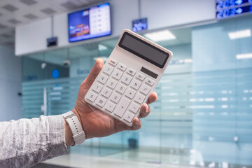 A hand holds a white calculator on a blurred background of a bank with exchange rates. Concept on the theme of business and financial literacy.