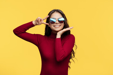 Smiling young lady in red dress and sun glasses is posing on yellow background. Showing peace gesture