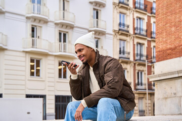 Young man sending a vocal message on his smartphone while sitting in the city