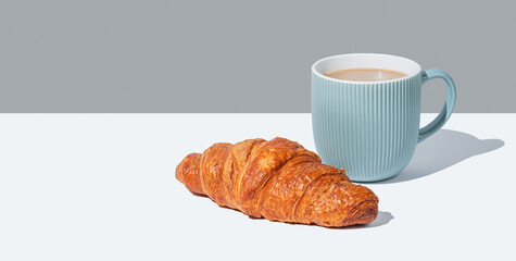 Fresh croissant and cup of coffee with milk on white and gray background with shadow.