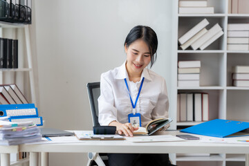 Focused Professional Woman: A smiling young woman in a crisp white shirt and lanyard sits at her...