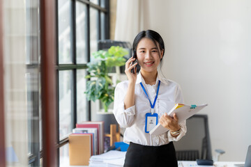 Confident Businesswoman on the Phone: A smiling young businesswoman, dressed in professional attire, confidently handles a phone call while holding documents.
