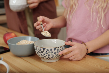 Side view close up of young girl adding oats to bowl in kitchen while preparing healthy breakfast with mother in kitchen copy space