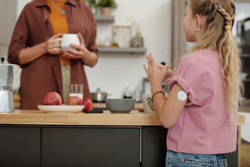 Back view of young girl with diabetes standing at kitchen counter in morning and chatting to mom...