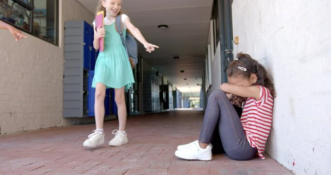 In school, girl pointing at another girl sitting on floor, feeling upset