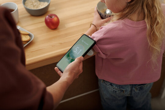 Close up of unrecognizable woman holding smartphone with green glucose reading to GCM device on daughters arm and caring for child with diabetes copy space 