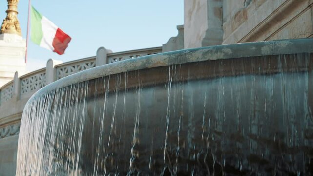 Slow-motion footage of a fountain's cascading water with the Italian flag waving in the background, filmed in Rome, evoking the city's rich heritage and iconic landmarks