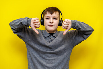 Ten year old boy in grey knitted sweater listening to music in black headphones and showing thumbs down gesture with both hands on yellow background.