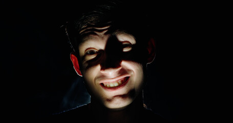 A man in the darkness with a twisted, unsettling grin, lit dramatically from below, casting eerie...