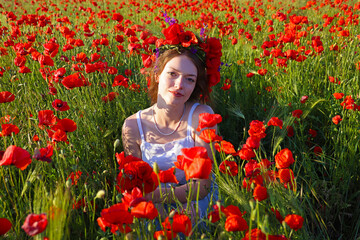 Portrait of a beautiful young girl with floral wreath on her head picking and enjoying in poppy field. Beauty in nature, rural landscape with blue sky, young girl and lots of red flowers in meadow