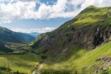 Serene Alpine valley with lush green hills, rocky cliffs, and a winding river under a partly cloudy sky. Tranquil and picturesque mountain scenery.