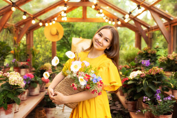 Portrait of a pretty teen girl in dress posing with bouquet of flowers in basket. Young girl holding flowers on backyard in wooden illuminated greenhouse with plants and flowers in pots	
